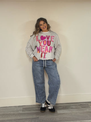 Person wearing a gray sweatshirt with colorful text and blue jeans against a plain background