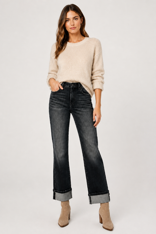 A woman modeling a pair of dark-wash cropped jeans with rolled cuffs, paired with a beige knit sweater and beige ankle boots, against a light gray studio background.