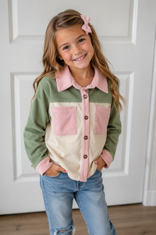 A young girl with a pink hair bow wearing a color-blocked button-up shirt with sage green, dusty pink, and cream panels, paired with blue jeans, standing in front of a white paneled door.