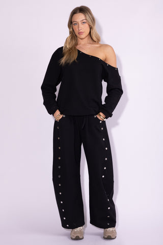 Woman wearing a black off-shoulder top and black pants with button details on a white background
