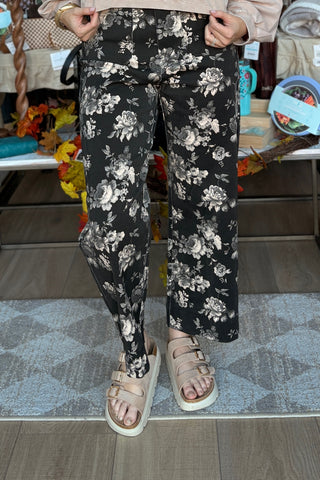 A person wearing black wide-leg floral pants with beige and gray rose patterns, paired with beige platform sandals, standing in a retail store setting.