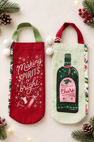 Two Christmas-themed wine bottle tote bags laid flat on a light beige background with pine branches, pinecones, and fairy lights. The left bag is red with green handles and white text reading 'Making Spirits Bright.