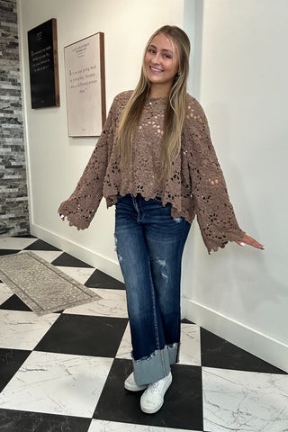 A woman modeling a brown lace long-sleeve crop top with scalloped edges, paired with blue jeans and white sneakers, standing in an indoor hallway with a black and white checkered floor.