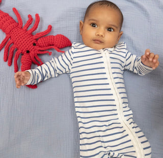 Baby in a striped onesie lying next to a red knitted lobster toy on a blue blanket.