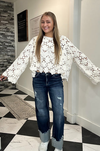 A young woman with blonde hair stands in a hallway wearing a white crochet long-sleeve top, dark blue jeans, and white sneakers.