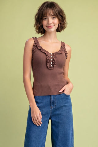 A woman modeling a brown ribbed sleeveless top with ruffled straps and a buttoned chest detail, paired with blue jeans, against a solid light olive-green background.