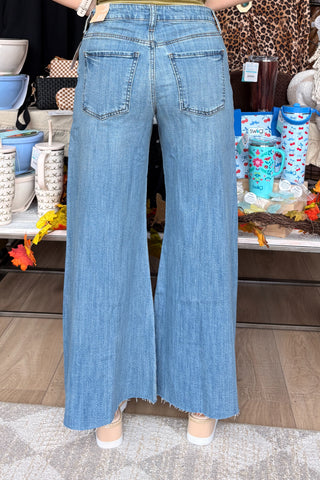 A person shown from the back wearing light blue wide-leg denim jeans with a frayed hem, standing in a retail store interior with shelves and bags in the background.