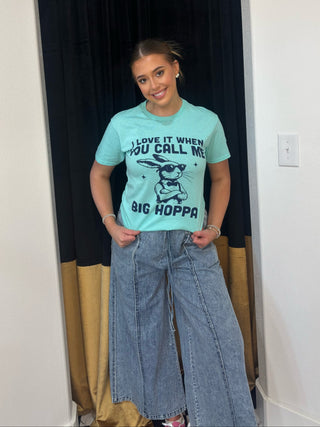 Person wearing a light blue t-shirt with text and graphics, standing in a room with a curtain and wall.