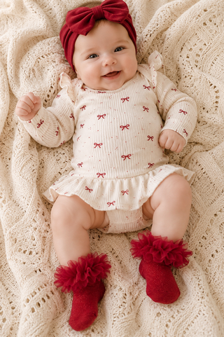 A baby lying on a cream knit blanket, wearing a cream ribbed long-sleeve romper with small red bow prints, a matching red bow headband, and red fuzzy booties.