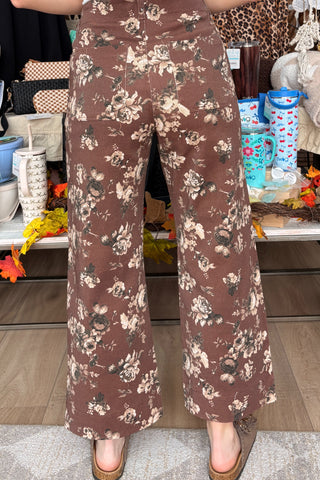 A person wearing brown wide-leg pants with a beige and white floral print, shown from the waist down in a retail store setting.