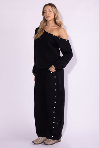 A woman modeling a black off-the-shoulder knit top and matching wide-leg pants with silver button details, standing against a light lavender studio background.