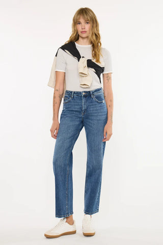 A model stands against a plain white studio background wearing a pair of high-waisted, straight-leg blue jeans, a white t-shirt, and white sneakers. A black and beige knit sweater is draped over her shoulders.