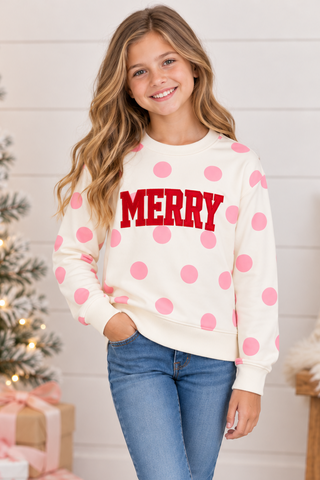 A young girl stands in a cozy holiday room wearing a cream sweatshirt with pink polka dots and a red 'MERRY' text graphic, paired with blue jeans and white sneakers.