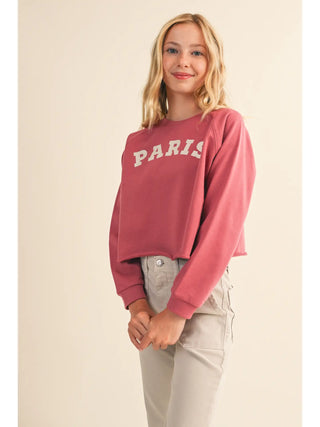 A young woman with blonde hair wearing a dusty rose pink cropped sweatshirt with 'PARIS' printed in white arched letters across the chest, paired with light beige pants, set against a solid cream background.