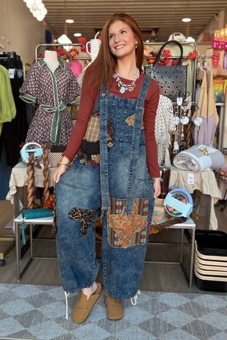 A woman stands in a clothing boutique wearing a blue denim overall-style jumpsuit with colorful patchwork pockets over a rust-brown long-sleeve top and tan slip-on shoes. Clothing racks and mannequins are visible in the background.