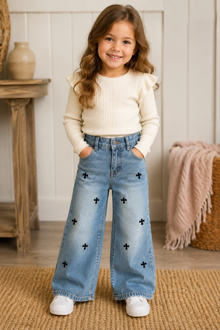 A young girl with long wavy brown hair stands smiling in a home setting, wearing light-wash blue wide-leg jeans with black cross details, a cream ribbed long-sleeve top, and white sneakers.
