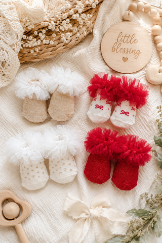 A styled flat-lay of baby booties and mittens arranged on a cream fabric background with decorative props. The set includes beige and white booties with fluffy white trim, white mittens with small red bows, and red booties with fluffy red trim.