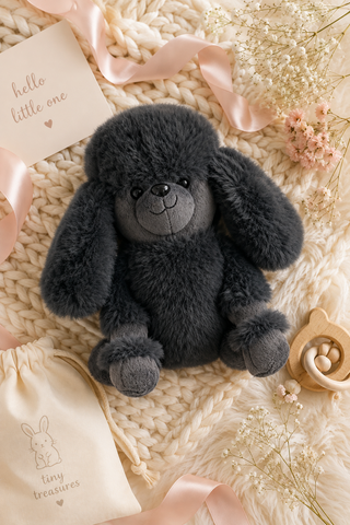 A dark gray plush puppy toy sits on a cream-colored chunky knit blanket, surrounded by baby-themed props including a cream drawstring pouch, a wooden pacifier clip, dried flowers, and a pink ribbon. A small card in the corner reads 'hello little one'.