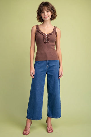 A woman modeling a brown sleeveless top with ruffle trim and button details, paired with blue wide-leg cropped jeans and pink sandals, against a solid light olive green background.