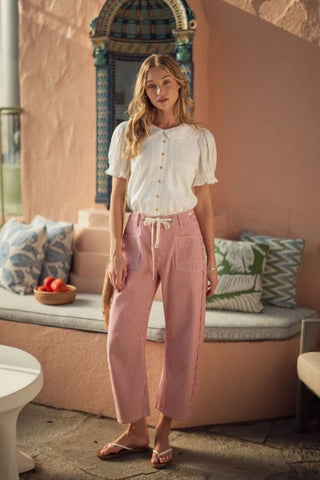 Woman in a white blouse and pink pants standing in front of a colorful wall with decorative pillows.