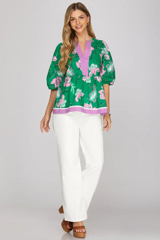 Woman wearing a green floral blouse with purple accents and white pants on a gray background