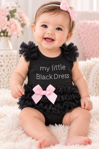 A baby girl wearing a black ruffled romper dress with 'my little Black Dress' printed on the front, sitting on a white fluffy rug.