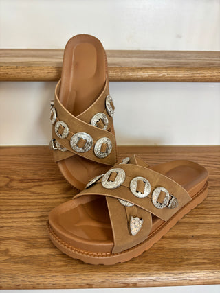 Pair of tan sandals with decorative buckles on a wooden surface.