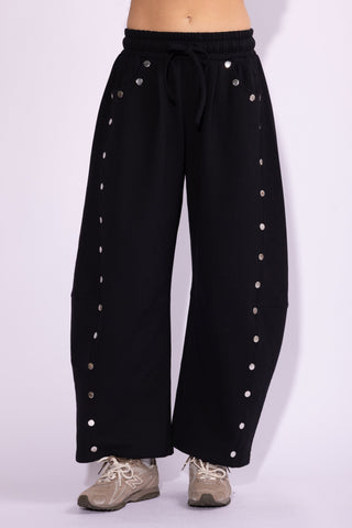 Black wide-leg sweatpants with silver metal studs down the sides and a drawstring waist, shown on a model from the waist down wearing beige sneakers against a light lavender studio background.
