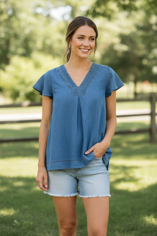 Woman wearing a blue top and denim shorts standing outdoors with greenery in the background