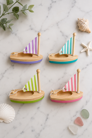 Four small wooden toy sailboats arranged on a white marble surface with decorative seashells and greenery. The boats have natural wood hulls and striped sails in purple, teal, lime green, and pink, with matching colored bottoms.