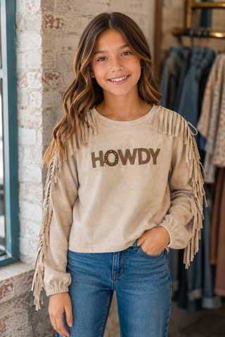 A young girl with long brown hair smiles while wearing a beige long-sleeve sweatshirt with fringe details and the word 'HOWDY' printed across the chest, paired with blue jeans in a boutique setting.