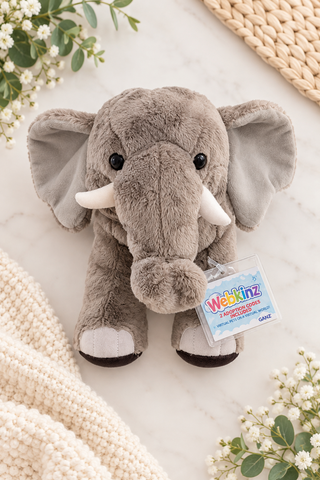 A plush gray elephant toy with white tusks and a hanging Webkinz tag, styled on a light marble surface with cream knit textiles and small white flowers.
