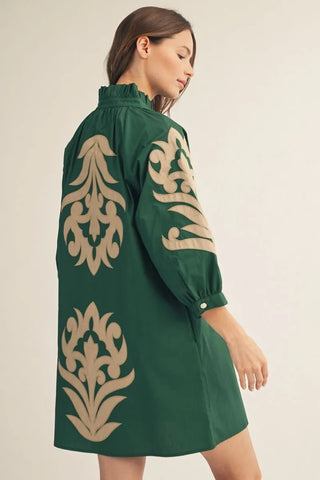 A woman wearing a dark green shift dress with large beige damask-style embroidery on the back and sleeve, shown from a side-back angle against a plain light beige studio background.