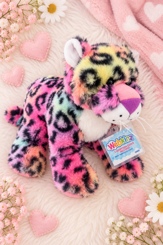 A colorful plush toy leopard with a rainbow gradient and black leopard spots, lying on a pink faux fur surface decorated with pink hearts, white daisies, and a pink blanket. A Webkinz tag is attached to its neck.