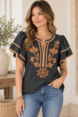 A woman wearing a black short-sleeve blouse with tan floral embroidery and trim, paired with blue jeans, standing in a softly lit indoor setting.