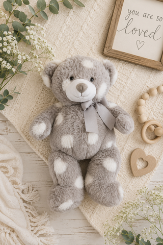 A plush gray teddy bear with white spots and a gray ribbon bow, styled on a cream knit blanket with baby props including a wooden heart, beads, eucalyptus, and a framed print reading 'you are so loved'.