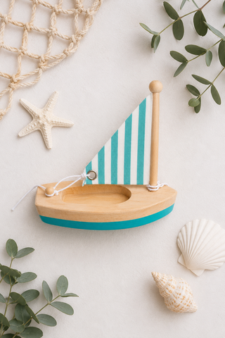 A wooden toy sailboat with a teal and white striped sail and a teal hull, styled on a light beige background with seashells, eucalyptus leaves, and a rope net.