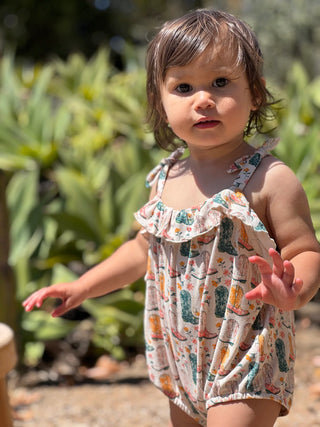 Child wearing a patterned romper standing outdoors with greenery in the background