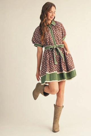 A woman modeling a short-sleeve mini dress with a brown and white floral pattern, green trim, and a matching green tie belt, paired with tan boots against a plain beige studio background.