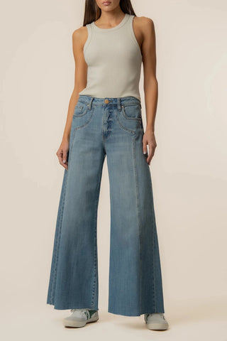 A model wearing a light beige ribbed tank top, wide-leg blue denim jeans, and light gray sneakers, standing against a solid warm beige studio background.