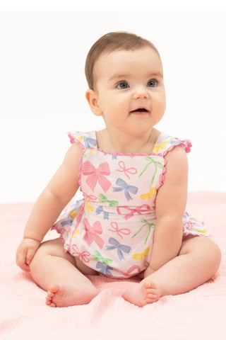 Baby sitting on a pink blanket wearing a colorful romper with bow patterns.