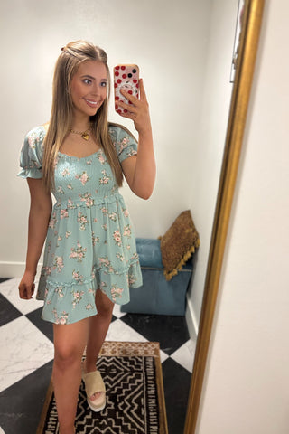 Woman taking a mirror selfie wearing a light blue floral dress in a room with a checkered floor.