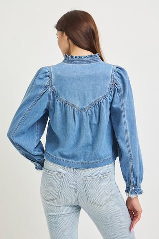 A woman shown from the back wearing a light blue denim jacket with long puff sleeves, a ruffled high neck, and gathered detailing across the upper back, paired with light blue patterned jeans against a plain light gray studio background.