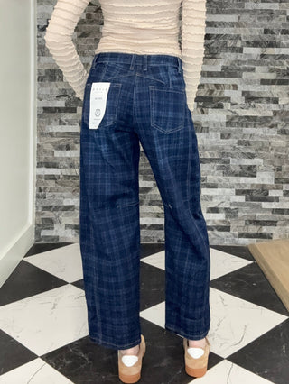 A woman shown from behind wearing a beige ribbed long-sleeve top and dark blue plaid wide-leg jeans, standing on a black and white checkered floor in front of a gray stone wall.