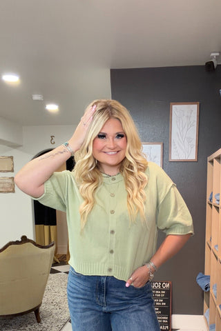 Woman in a green shirt and blue jeans standing in a room with checkered floor and wooden shelves.