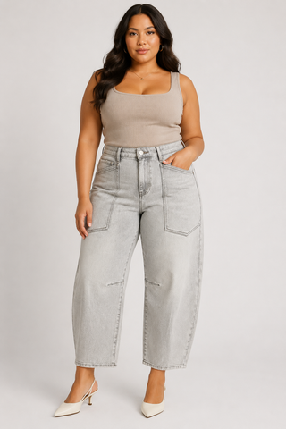 A woman modeling light-wash wide-leg cropped jeans with a beige ribbed tank top and white pointed-toe heels, standing against a light gray studio background.