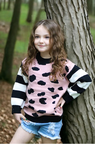 Young girl in a pink sweater with black spots and denim shorts standing next to a tree in a forest.