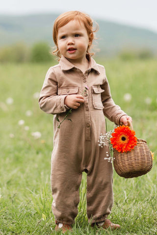 Child in a brown outfit holding flowers and a basket in a grassy field