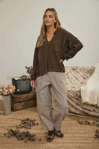A woman stands in a styled indoor room wearing a dark brown long-sleeve V-neck top and beige-and-gray vertically striped pants, with brown slip-on shoes. The background includes a vintage television on a crate, a floral-covered sofa, woven rug, and scattered dried flowers.