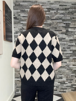 A person viewed from behind wearing a short-sleeve knit sweater with a black and off-white harlequin diamond pattern, paired with black pants, standing in a hallway with a gray stone accent wall.
